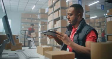 Afro-Amerikalı depo görevlisi paketleri tablet bilgisayar kullanarak tarıyor, postanede çalışıyor. Nakliye için karton kutular taşıyan tasnif merkezi çalışanları. Çevrimiçi mağaza ürünleriyle lojistik depolama.