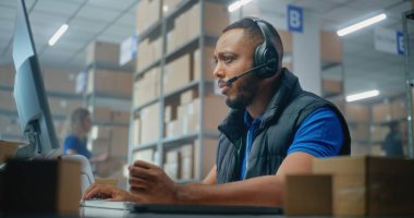 Afrikalı Amerikalı lojistik müşteri destek uzmanı müşteriyle kulaklık görüşmeleri yapıyor, bilgisayar kullanıyor, posta servisinde çalışıyor. Teslimat için kutu taşıyan tasnif merkezi çalışanları. Çevrimiçi depo.