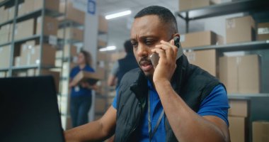 E-ticaret mağazası veya postanenin depo perakende satış tesisi: Afrikalı Amerikalı lojistik koordinatörü telefonla konuşuyor, dizüstü bilgisayar kullanıyor. Teslimat için karton kutular taşıyan tasnif merkezi çalışanları.