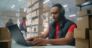 Kulaklıkla Afrikalı Amerikalı lojistik koordinatörünün portresi müşteriyle konuşuyor, posta servisinde ya da çevrimiçi depolamada dizüstü bilgisayarla çalışıyor, kameraya bakıyor. Sıralama merkezi çalışanları kutucukları işaretliyor