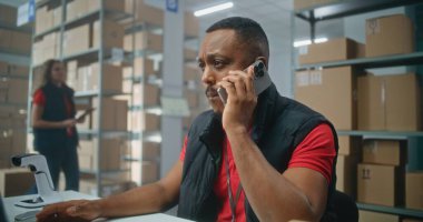 E-ticaret veya posta servisinin depo perakende tesisi: Afrikalı Amerikalı lojistik koordinatörü telefonla konuşuyor, bilgisayar kullanıyor. Teslimat için karton kutuları kontrol eden sıralama merkezi çalışanları.