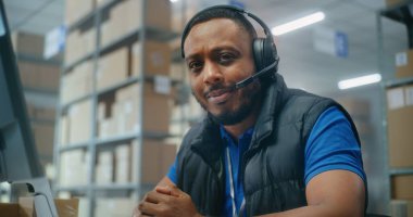 Afrika kökenli Amerikalı müşteri destek uzmanı müşteriyle kulaklık konuşmaları yapıyor, bilgisayar kullanıyor, kameraya bakıyor. Kutu taşıyan tasnif merkezi çalışanları. Posta servisi veya çevrimiçi depo.
