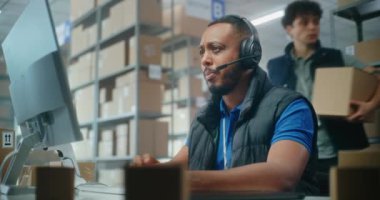 Afrika kökenli Amerikalı müşteri destek uzmanı müşteriyle kulaklık konuşmaları yapıyor, bilgisayar kullanıyor, kameraya bakıyor. Kutu taşıyan tasnif merkezi çalışanları. Posta servisi veya çevrimiçi depo.