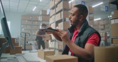 Afro-Amerikalı depo görevlisi paketleri tablet bilgisayar kullanarak tarıyor, postanede çalışıyor. Nakliye için karton kutular taşıyan tasnif merkezi çalışanları. Çevrimiçi mağaza ürünleriyle lojistik depolama.