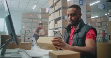 Afro-Amerikan lojistik müdürü akıllı telefon kullanarak paketleri tarıyor, posta servisi deposunda çalışıyor. Nakliye için karton kutular taşıyan tasnif merkezi çalışanları. E-ticaret emirleriyle birlikte depolama.