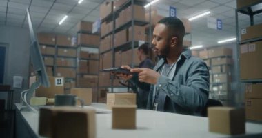 Afro-Amerikan depo çalışanı paketleri tablet bilgisayar kullanarak tarıyor, bilgisayar ekranına bakıyor, postanede çalışıyor. Teslimat için kutu taşıyan tasnif merkezi çalışanı. E-ticaret mağazasının deposu.