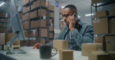 Sıralama merkezi: Erkek Afro-Amerikan depo çalışanı müşteriyle cep telefonuyla konuşur, envanteri bilgisayarda kontrol eder, teslimat için paketleri hazırlar. Çevrimiçi E-ticaret mağazasının siparişleriyle birlikte depolama