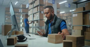 Afro-Amerikan lojistik müdürü akıllı telefon kullanarak paketleri tarıyor, posta servisi deposunda çalışıyor. Merkez çalışanları karton kutuları nakliye için kontrol ediyor. E-ticaret emirleriyle birlikte depolama.