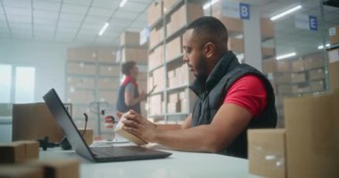 Afro-Amerikan postane çalışanlarının portresi barkod tarayıcısıyla tarama yapıyor, dizüstü bilgisayar kullanıyor, kameraya bakıyor. Teslimat için merkez çalışanları kutuları kontrol ediyor. Çevrimiçi mağaza deposu.