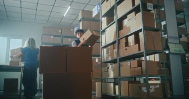 Erkek depo çalışanının karton paketi raftan alıp müşterilere göndermek için palet kamyona yüklemesi. Arka planda çalışan tasnif merkezi çalışanları. Teslimat servisi.