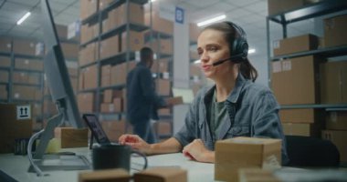 Posta servisi deposu veya E-ticaret mağazası: Müşteriyle kulaklıkla kadın lojistik koordinatörü konuşuyor, bilgisayar kullanıyor. İşçi sınıflandırma merkezi müşterilere karton kutular gönderme.