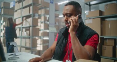 E-ticaret veya posta servisinin depo perakende tesisi: Afrikalı Amerikalı lojistik koordinatörü telefonla konuşuyor, bilgisayar kullanıyor. Teslimat için karton kutuları kontrol eden sıralama merkezi çalışanları.