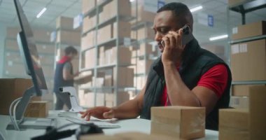 Postane veya E-ticaret mağazası depolama tesisi: Cep telefonuyla konuşan Afrikalı Amerikalı lojistik koordinatörü, bilgisayar kullanıyor. Merkez çalışanları nakliye için karton kutuları kontrol ediyor.