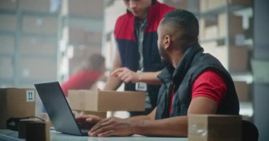 Afro-Amerikan lojistik uzmanı postanedeki dizüstü bilgisayarda çalışıyor. Depo çalışanı teslimat bilgilerini kontrol etmek için paket getiriyor. E-ticaret çevrimiçi mağazasının sıralama merkezi. Yavaş çekim.