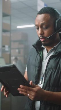 Afro-Amerikalı Müşteri Destek Uzmanı Kulaklık Görüşmeleri, Tablet Bilgisayar Kullanıyor. Depo çalışanı teslimat için kutuları hazırlıyor. E-Ticaret Mağaza Sıralama Merkezi. Dikey Çekim