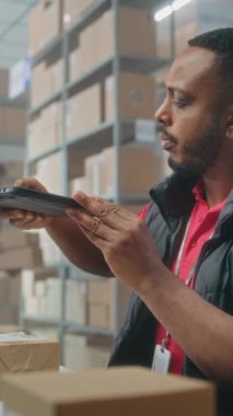 Afro-Amerikalı depo görevlisi paketleri tablet bilgisayar kullanarak tarıyor, postanede çalışıyor. Nakliye için karton kutular taşıyan tasnif merkezi çalışanları. Çevrimiçi mağaza lojistik deposu. Dikey çekim