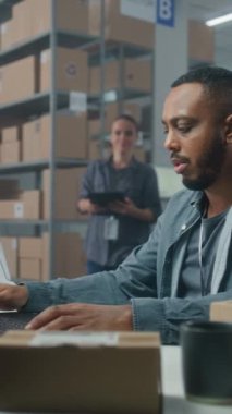 Depo tesisi, Afrikalı Amerikalı lojistik uzmanı dizüstü bilgisayardaki envanteri analiz ediyor, iş arkadaşıyla konuşuyor. Kadın depo çalışanı tablet bilgisayar kullanıyor, teslimat için paketleri kontrol ediyor. Dikey çekim.