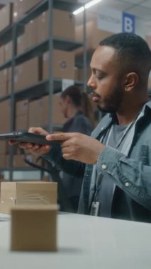 Afro-Amerikan depo ortağı paketleri tablet bilgisayar kullanarak tarıyor, bilgisayar ekranına bakıyor, E-ticaret mağazasının deposunda çalışıyor. Teslimat için kutu taşıyan tasnif merkezi çalışanı. Dikey çekim.