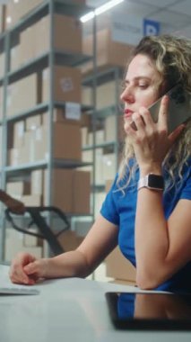 Postanenin depo tesisi, E-ticaret mağazası: Kadın lojistik koordinatörü cep telefonuyla konuşuyor, bilgisayar kullanıyor. Teslimat merkezindeki çalışanlar müşterilere kargo için kutu taşıyorlar. Dikey çekim