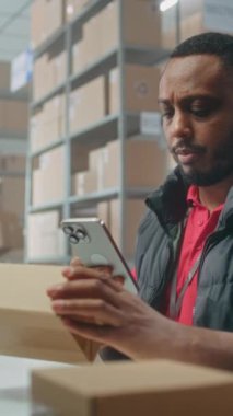 Afro-Amerikan lojistik müdürü akıllı telefon kullanarak paketleri tarıyor, depoda çalışıyor. Nakliye için karton kutular taşıyan tasnif merkezi çalışanları. E-ticaret emirleriyle dolu bir depo. Dikey video.