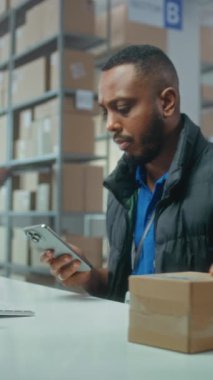 Afro-Amerikan lojistik müdürü akıllı telefon kullanarak paketleri tarıyor, posta servisi deposunda çalışıyor. Tasnif merkezi çalışanları nakliye için kutuları işaretliyor. E-ticaret emirleriyle dolu bir depo. Dikey çekim.