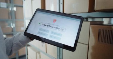 Posta Deposu İşçisi, Karton Kutudaki QR kodunu barkod tarayıcı ile tarıyor, Paket Bilgilerini izliyor. Tablet Bilgisayar Ekranı Paket Yönlendirmesini Kontrol Etmek için Sevkiyat İzleme Yazılımını gösterir. Kapat..