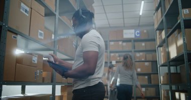 Yüksek Teknolojik Modern Depo: Afro-Amerikan Envanter Uzmanı Kulaklık Tarayıcı Paketleri Giyiyor, Tablet Bilgisayar Kullanıyor. Gelişmiş Tam Vücut Robotik Dış iskelet İşçisi Ağır Karton Kutuyla Yürüyor.