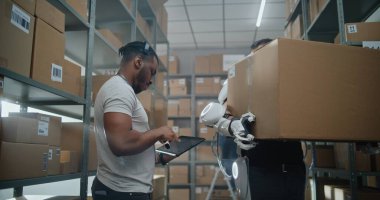 Yüksek Teknolojik Gelişmiş Depo: Afro-Amerikan Yönetici Kulaklık Tarama Paketleri Giyiyor, Tablet Bilgisayar Envanterini Kontrol Ediyor. Fütürist Full Body Exoskeleton Walks 'ta Karton Kutuyla Yürüyen İşçi.