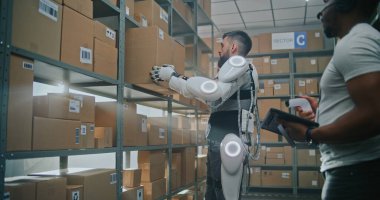 High-Tech Modern Warehouse: Worker Wearing Futuristic Full Body Robotic Exoskeleton Puts Cardboard Box on Shelf. African American Logistics Specialist Scans Parcel, Checks Inventory on Tablet Computer clipart