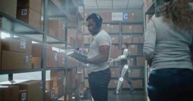 Yüksek Teknolojik Gelişmiş Depo: Afro-Amerikan Envanter Uzmanı Kulaklık, Tarama Paketleri, Tablet Bilgisayar Kullanıyor. Arkaplanda Karton Yürüyüşlü Robotik Dış iskelet İşçisi.
