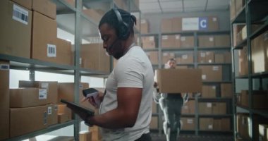 Yüksek Teknolojik Modern Depo: Afro-Amerikan Envanter Uzmanı Kulaklık Tarayıcı Paketleri Giyiyor, Tablet Bilgisayar Kullanıyor. Gelişmiş Tam Vücut Robotik Dış iskelet İşçisi Ağır Karton Kutuyla Yürüyor.