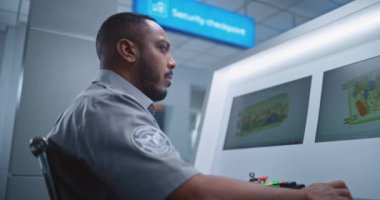Afrikalı Amerikalı Güvenlik Subayı Modern Yazılım kullanarak Bilgisayar Monitörlerinde Bavul Gösterimini Denetliyor. Uçak Uçuşu için havaalanı güvenlik kontrol noktasındaki konveyör bandında gelişmiş X-ray taraması. Kapat..