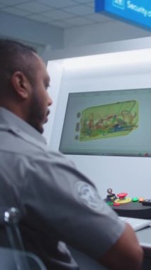 Havaalanı Güvenlik Kontrol Noktası: Afro-Amerikan Güvenlik Subayı Bilgisayar ekranlarında bagaj taramasını izliyor, Bavulda Şüpheli Nesne Buluyor. Gelişmiş X-ray Tarama Teknolojisi. Dikey Çekim.