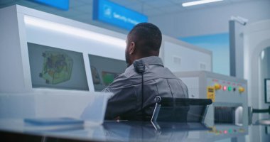 Havaalanı Güvenlik Kontrol Noktası: Afrikalı Amerikan Güvenlik Subayı Bilgisayar ekranlarında bagaj taramasını izliyor. Ön planda, Yolcular Uçak Uçuşundan Önce Kişisel Eşyaları Tablalara Koyuyor.