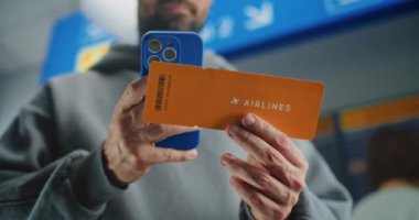 Uluslararası Havaalanı Terminali: Uçuş Bilgilerini Kontrol Etmek için Akıllı Telefon kullanarak İnsan Tarama Uçak Biletini Kapatın. Gezgin Seyahate Çıkıyor. Arka planda Yolcular ve Varış Ekranı.