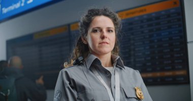 Havaalanı Terminalindeki güvenlik görevlisinin portresi: Üniformalı kadın TSA çalışanı kameraya bakıyor. Arkaplanda Geliş ve Gidiş Bilgileri Bilgilerine Bakan Çeşitli Yolcu topluluğu.