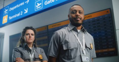 Uluslararası Havalimanı Terminalindeki Profesyonel TSA çalışanlarının Portresi: Kameraya Bakan Üniformalı İki Güvenlik Memuru. Arka planda Dijital Uçuş Bilgileri Ekranı. Doli Vuruşu.