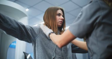 Havaalanı Güvenlik Kontrol Noktası: Kadın Güvenlik Görevlisi, X-ray Metal Dedektörü Kapıları Taradıktan sonra Genç Kadın Bedenini Şüpheli Eşyalar İçin Denetliyor. TSA tarama prosedürleri. Turist Tatile Çıkıyor.