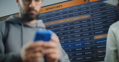 Uluslararası Havalimanı Terminali: Uçağa biniş telefonu kullanan adam bilgileri kontrol ediyor. Arkaplanda Dijital Geliş ve Gidiş Bilgileri Görünümü. Gezgin Tatile Çıkıyor.