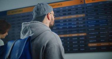 Kalabalık Havaalanı Terminali: Gelen ve Giden Uçuş Bilgilerini Arayan Sırt çantalı Adam, Biniş Uçağı Bilgilerini Kontrol Etmek için Akıllı Telefon 'u kullanıyor. Turist Tatile Çıkıyor.