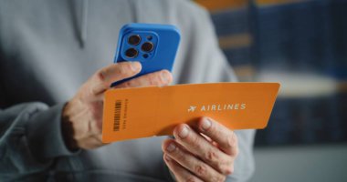 Uluslararası Havaalanı Terminali: Uçuş Bilgilerini Kontrol Etmek için Akıllı Telefon kullanarak Erkek Yolcu Tarama Biletini Kapatın. Gezgin Seyahate Çıkıyor. Arkaplanda Dijital Ayrılış Tahtası.