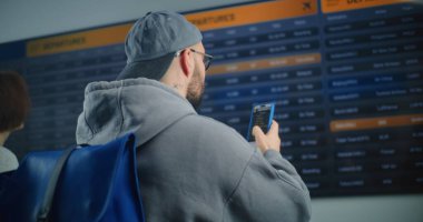 Kalabalık Havaalanı Terminali: Gelen ve Giden Uçuş Bilgilerini Arayan Sırt çantalı Adam, Biniş Uçağı Bilgilerini Kontrol Etmek için Akıllı Telefon 'u kullanıyor. Turist Tatile Çıkıyor.