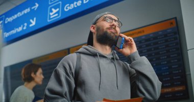 Havaalanı Terminali: Erkek Yolcu Tutma Uçak Bileti, Uçuş Bilgilerini Kontrol Etmek için Telefonla Konuşuyor. Arkaplanda İnsanlar ve Dijital Geliş ve Gidiş Bilgi Ekranı. Turist Seyahate Çıktı.