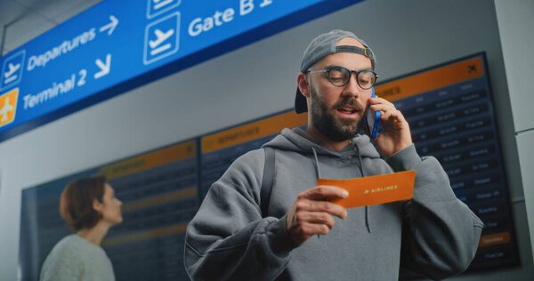 Airport Terminal: Male Passenger Holding Plane Ticket, Talking by Phone to Check Flight Information. People and Digital Arrival and Departure Information Screen in Background. Tourist Going on Trip.