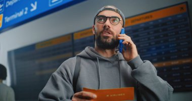 Havaalanı Terminali: Erkek Yolcu Tutma Uçak Bileti, Uçuş Bilgilerini Kontrol Etmek için Telefonla Konuşuyor. Arkaplanda İnsanlar ve Dijital Geliş ve Gidiş Bilgi Ekranı. Turist Seyahate Çıktı.