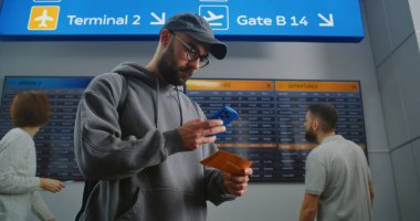 Yoğun Havaalanı Terminali: Sırt çantalı adam Uçuş Bilgi Tarama Biletini Telefon Kullanarak Kontrol Ediyor. Gezgin Tatile Çıkıyor. Arka plandaki Yolcular için Dijital Geliş ve Gidiş Kurulu.