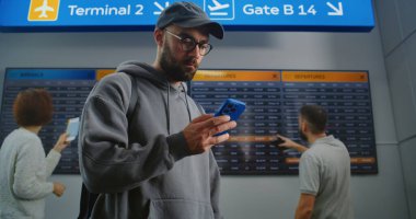 Yoğun Havaalanı Terminali: Sırt çantalı adam Uçuş Bilgi Tarama Biletini Telefon Kullanarak Kontrol Ediyor. Gezgin Tatile Çıkıyor. Arka plandaki Yolcular için Dijital Geliş ve Gidiş Kurulu.