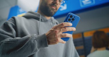 Uluslararası Havaalanı Terminali: Uçuş Bilgilerini Kontrol Etmek için Akıllı Telefon kullanarak İnsan Tarama Uçak Biletini Kapatın. Gezgin Seyahate Çıkıyor. Arka planda Yolcular ve Varış Ekranı.