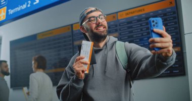 Kalabalık Havalimanı Terminali: Mutlu Erkek Yolcu Bekleme Uçağı Biletleri, Smartphone kullanarak Selfie çekmek, Kameraya Gülümsemek. Arka planda Dijital Uçuş Bilgileri Ekranı. Gezgin Seyahate Çıkıyor.