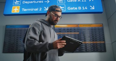 Kalabalık Havaalanı Terminali: Sırt çantalı adam Tablet Bilgisayar kullanıyor, Dijital Uçuş Bilgi Ekranından Gidişini Denetler. Turist Tatile Gidiyor. Arkaplanda Havalimanı Zaman Çizelgesi Görüntüsü.